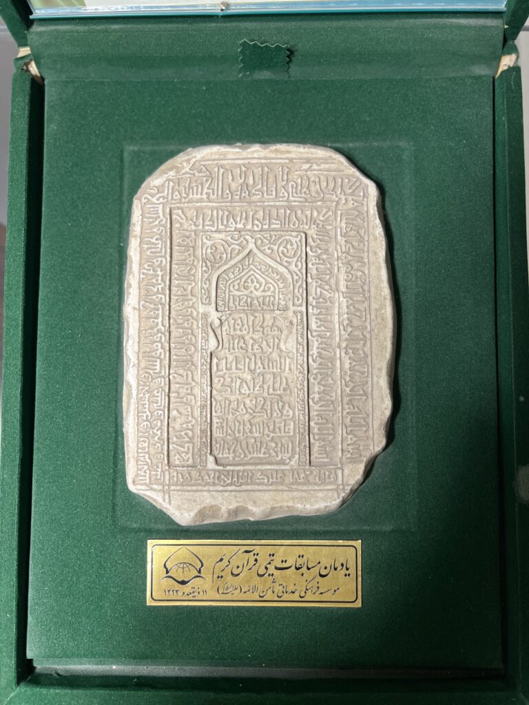 «تصویر سنگ تاریخی منقوش با خطوط اسلامی، منسوب به قدیمی‌ترین سنگ مزار حضرت امام رضا (ع)، نگهداری‌شده در قاب مخمل سبز با پلاک معرفی آستان قدس رضوی